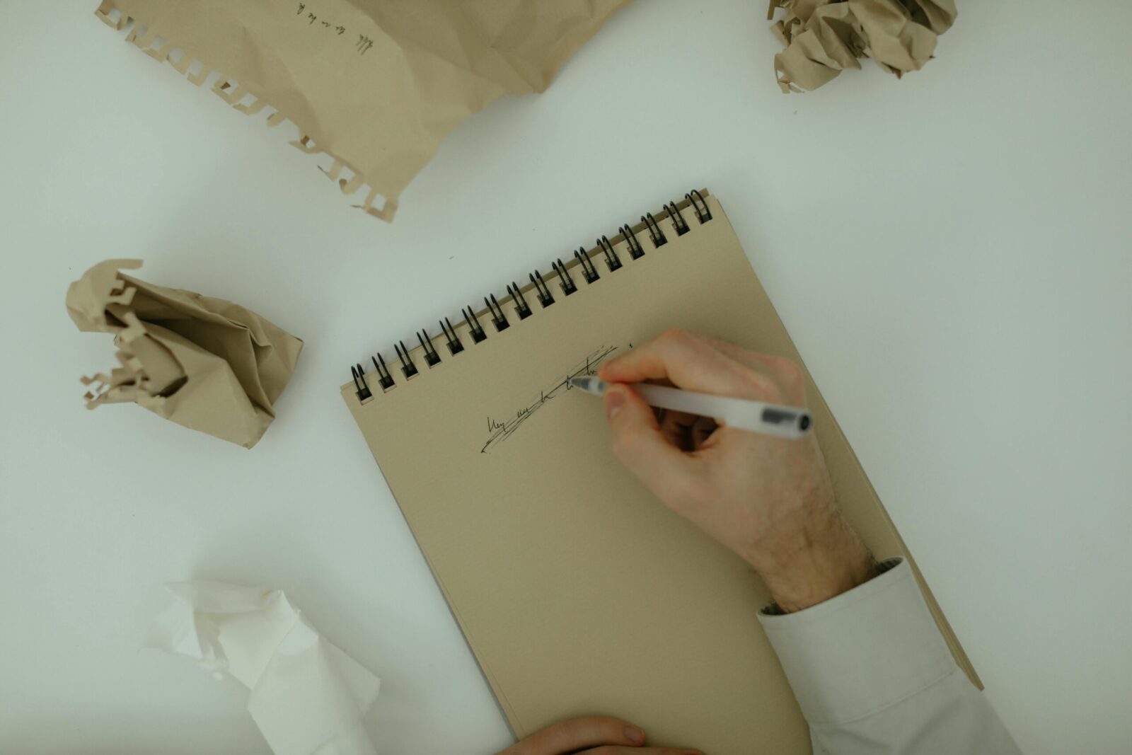 Person writing on a notepad surrounded by crumpled paper, symbolizing brainstorming and creativity.
